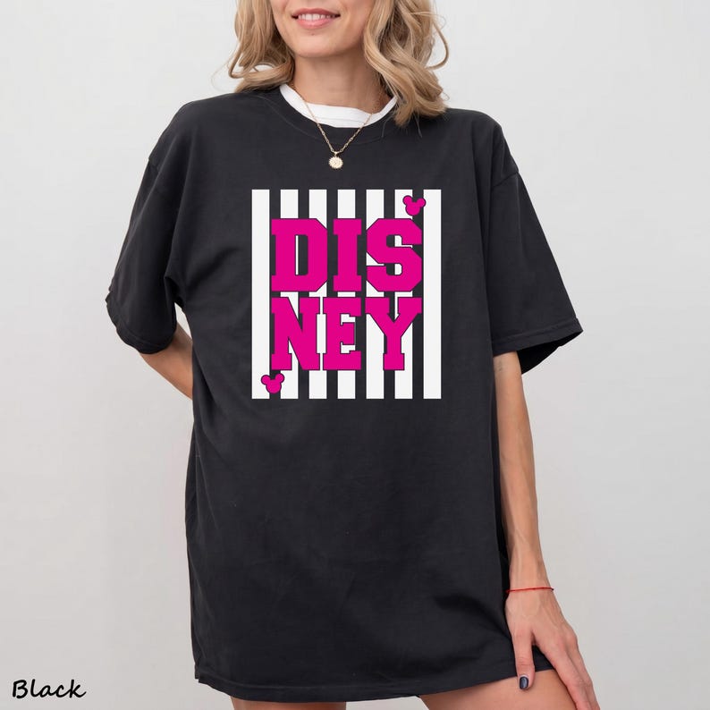 May include: Black t-shirt with the word "DISNEY" in bright pink block letters, set against a white and black striped background. Small pink hearts are placed around the word. The word "Black" is printed at the bottom left.