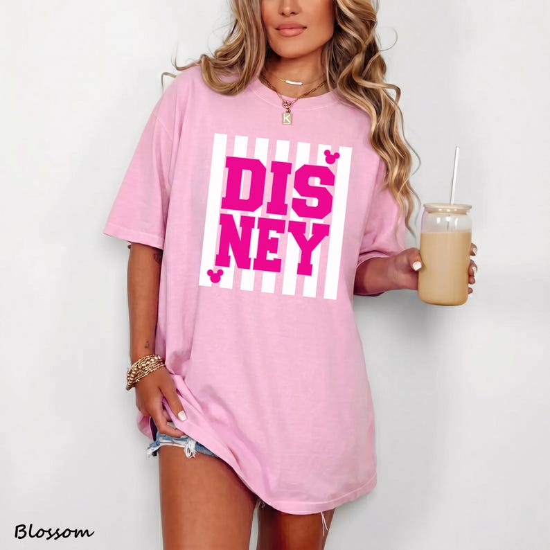 May include: A light pink t-shirt with the word "DISNEY" in bright pink block letters, set against a white striped background. The shirt has short sleeves and a relaxed fit. A clear glass with a straw is held in the hand.