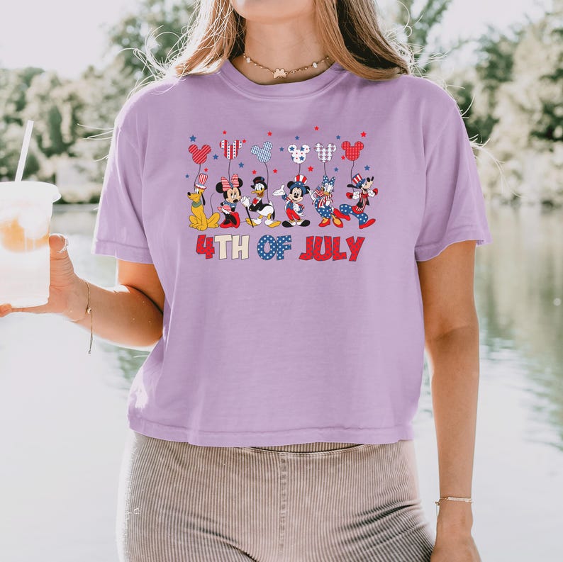 May include: A light purple t-shirt featuring a graphic of Disney characters including Mickey Mouse, Minnie Mouse, Donald Duck, and Pluto dressed in patriotic red, white, and blue outfits, holding balloons, with the text 4TH OF JULY printed below.