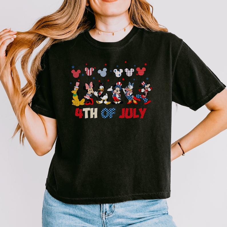 May include: A black short-sleeved t-shirt featuring a graphic of six cartoon characters dressed in red, white, and blue patriotic outfits. Above the characters are six Mickey Mouse ear silhouettes. The text 4TH OF JULY is printed below.