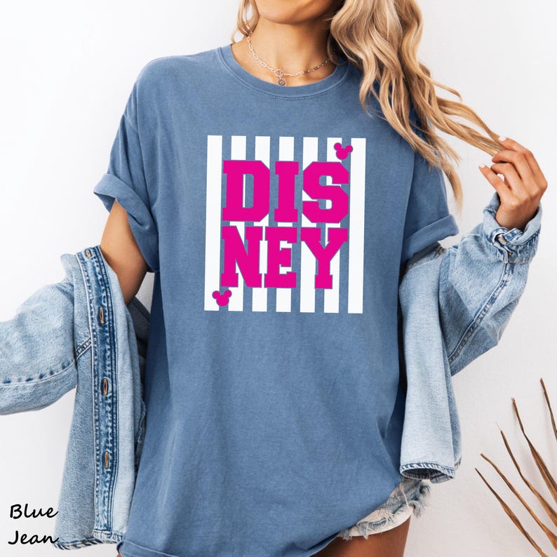 May include: Blue jean-colored t-shirt with the word "DISNEY" in bright pink letters, set within a white striped rectangle. Small pink Mickey Mouse head icons are placed near the letters. The shirt has rolled-up sleeves.