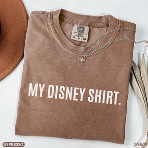 May include: Espresso-colored t-shirt with the text "MY DISNEY SHIRT." A silver necklace with a pendant rests on the shirt. The shirt is a Comfort Colors brand, size S-P-CH.