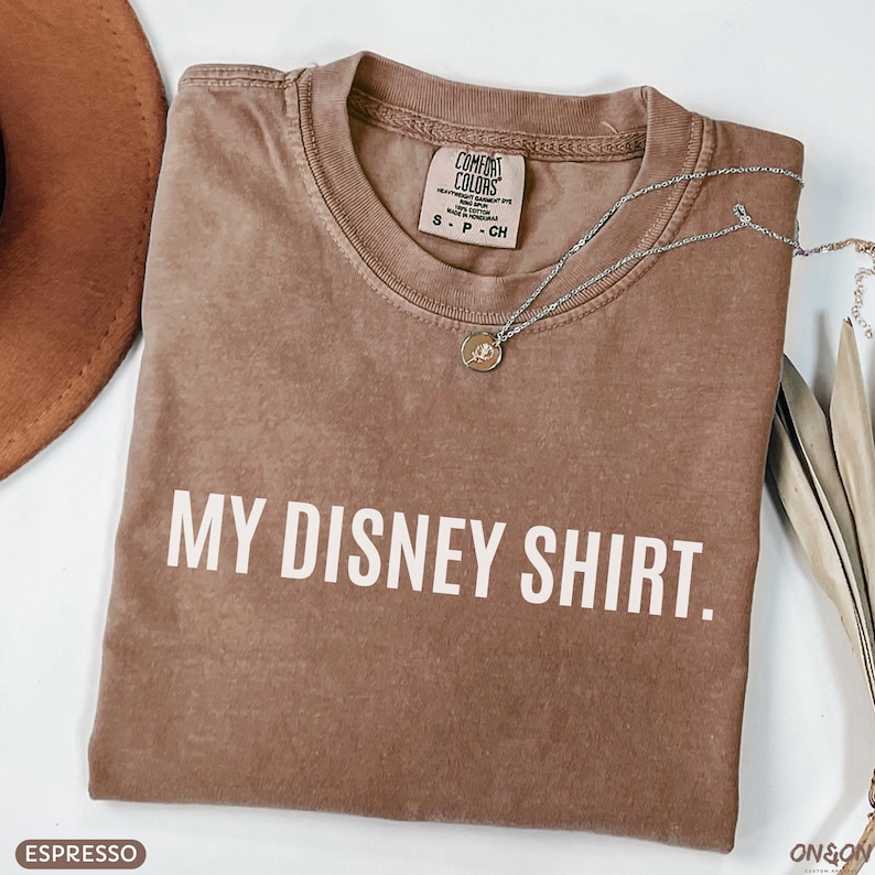May include: Espresso-colored t-shirt with the text "MY DISNEY SHIRT." A silver necklace with a pendant rests on the shirt. The shirt is a Comfort Colors brand, size S-P-CH.