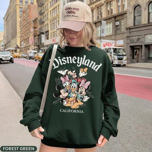 May include: Forest green Disneyland sweatshirt featuring Mickey Mouse, Minnie Mouse, Donald Duck, Daisy Duck, Goofy, and Pluto. The word "Disneyland" is arched across the top in white script, with "California" below. The person is wearing a tan baseball cap.