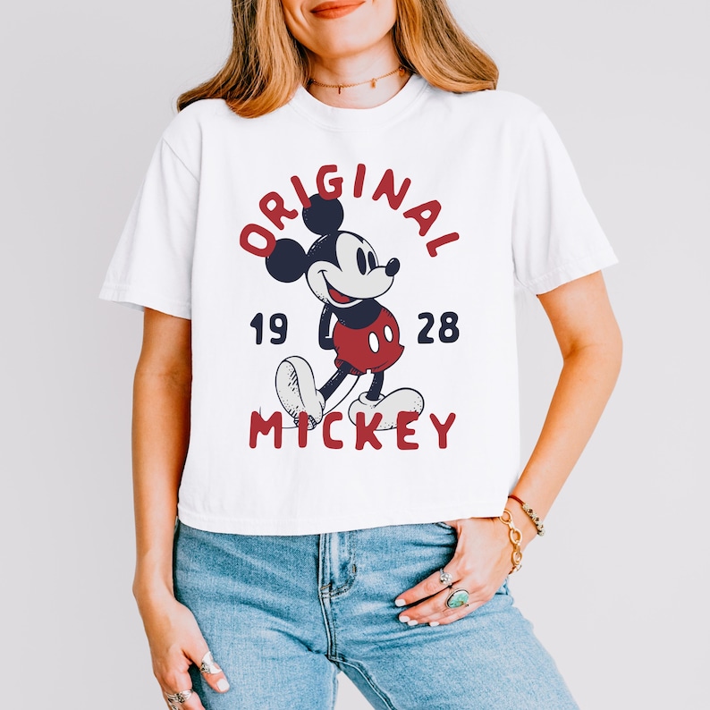 May include: A white short-sleeved t-shirt featuring a graphic of Mickey Mouse in the center. The text reads ORIGINAL above the character, 19 28 on the sides, and MICKEY below in red and dark blue lettering. The design has a distressed texture.