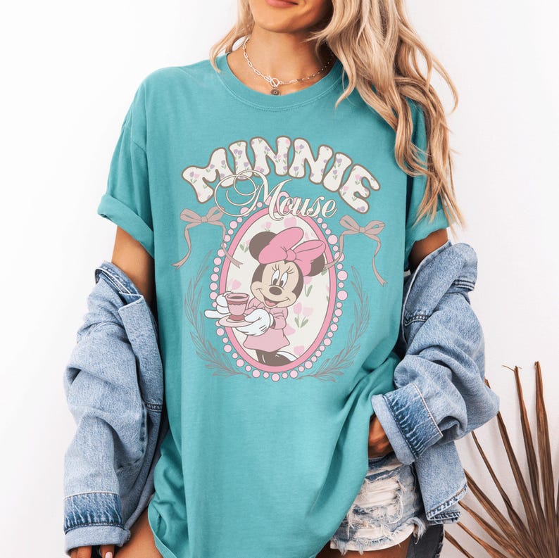 May include: A teal short-sleeve t-shirt featuring a graphic of Minnie Mouse holding a teacup inside an oval frame. The text MINNIE MOUSE is printed above the character in a decorative font, accented by pink bows and a floral pattern background.