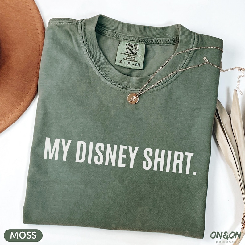 May include: A moss green t-shirt with the text "MY DISNEY SHIRT." printed in white. The shirt is folded and has a small tag at the neckline. A gold necklace with a pendant rests on the shirt. A brown hat is partially visible.