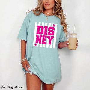 May include: A light blue t-shirt with the word "DISNEY" in bright pink, set within a white striped rectangle. The shirt has short sleeves and a relaxed fit. The text "Chalky Mint" is at the bottom left.