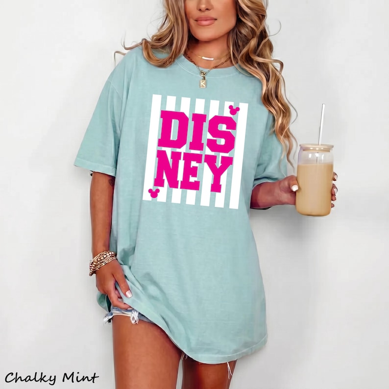 May include: A light blue t-shirt with the word "DISNEY" in bright pink, set within a white striped rectangle. The shirt has short sleeves and a relaxed fit. The text "Chalky Mint" is at the bottom left.