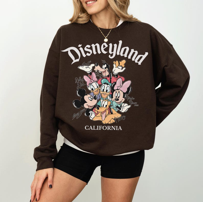 May include: A brown Disneyland sweatshirt featuring a graphic of Mickey Mouse, Minnie Mouse, Donald Duck, Daisy Duck, Goofy, and Pluto. The word "Disneyland" is arched across the top in white, with "CALIFORNIA" below.