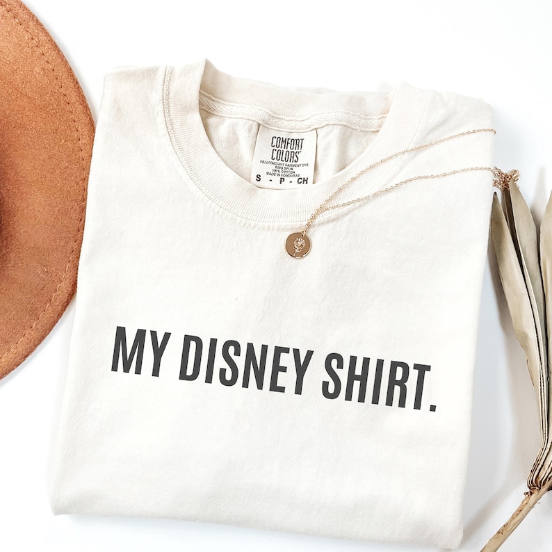May include: Cream-colored t-shirt with the text "MY DISNEY SHIRT." in black, featuring a gold necklace with a pendant. The shirt is from Comfort Colors and is laid flat on a white surface.