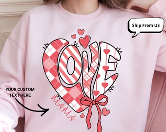 May include: A light pink sweatshirt with a large heart-shaped graphic. The graphic features the word "LOVE" in a stylized font, with red and white patterns and small hearts. The word "MAMA" is written below the heart.