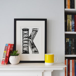 May include: Black and white illustration of the letter K, formed by the city of Krakow, Poland. The illustration is framed and displayed on a white shelf with a yellow mug and books.