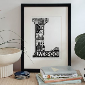 May include: Black and white illustration of the letter L made up of iconic landmarks and symbols from Liverpool, England. The letter is framed in a black frame and is displayed on a table with books.