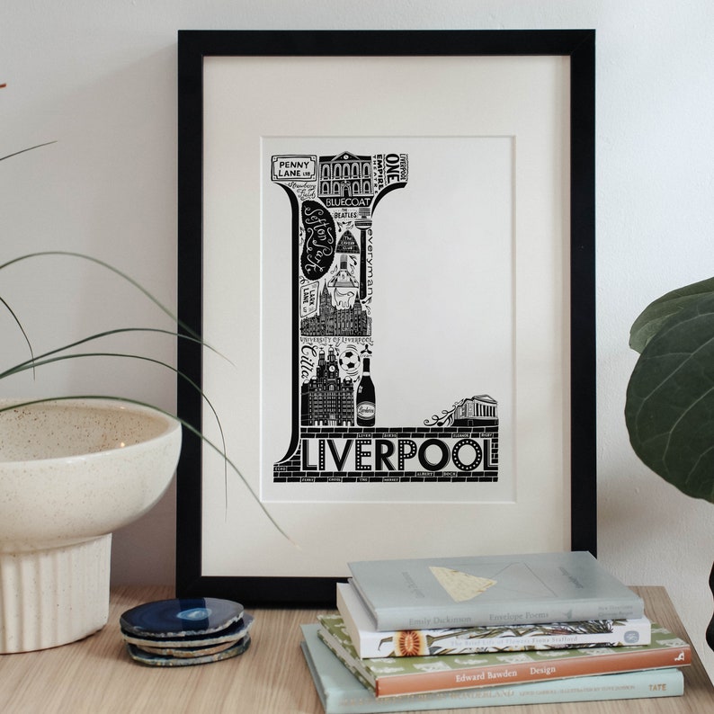 May include: Black and white illustration of the letter L made up of iconic landmarks and symbols from Liverpool, England. The letter is framed in a black frame and is displayed on a table with books.