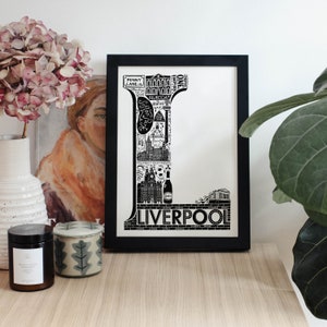 May include: Black and white illustration of the letter L made up of iconic landmarks and symbols from Liverpool, England. The letter is framed in a black frame and is displayed on a wooden surface.