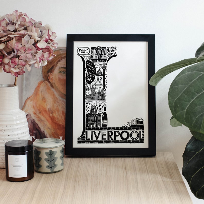 May include: Black and white illustration of the letter L made up of iconic landmarks and symbols from Liverpool, England. The letter is framed in a black frame and is displayed on a wooden surface.