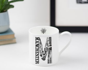 May include: White ceramic mug with a black and white illustration of the city of Manchester, England. The illustration is in the shape of the letter "M" and includes landmarks such as the Manchester Arndale, the Manchester Town Hall, and the Manchester Bee. The mug has a black handle.