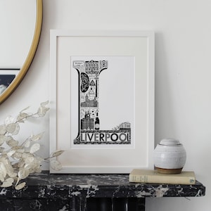 May include: Black and white illustration of the letter L made up of iconic landmarks and buildings from Liverpool, England. The word "Liverpool" is printed below the letter.