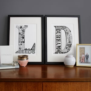 May include: Two black framed prints of the letters "L" and "D" made up of illustrations of landmarks in Leeds and Durham, England. The prints are on a wooden table with a small white ceramic jar, a pink ceramic jar, and a gold framed photo of a couple.