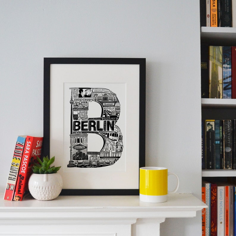 May include: Black and white print of the letter "B" made up of iconic landmarks and images from Berlin, Germany. The print is framed in a black frame and is displayed on a white mantel.