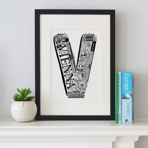 May include: Black and white illustration of the letter V made up of iconic landmarks and symbols from Vienna, Austria. The letter is framed in a black frame and displayed on a white shelf with travel books.
