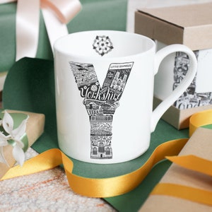 May include: White ceramic mug featuring a black and white illustrated "Y" filled with Yorkshire landmarks and the word "Yorkshire". The mug has a handle and a floral design inside the rim. Gift-wrapped presents and ribbons surround the mug.