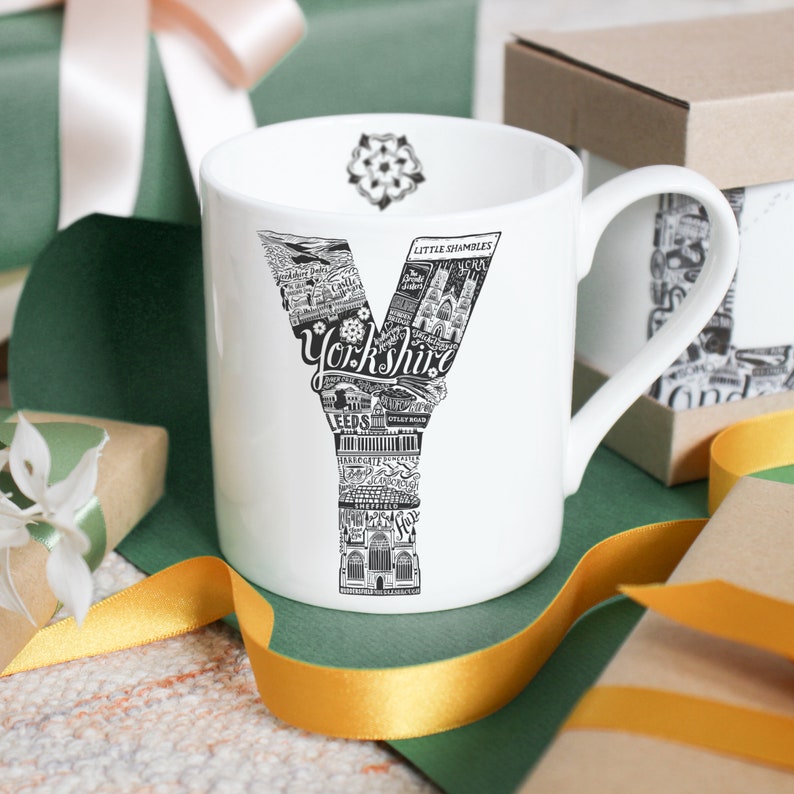 May include: White ceramic mug featuring a black and white illustrated "Y" filled with Yorkshire landmarks and the word "Yorkshire". The mug has a handle and a floral design inside the rim. Gift-wrapped presents and ribbons surround the mug.