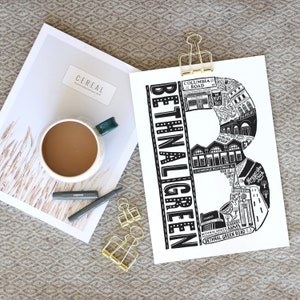 May include: A black and white print of the letter "B" filled with illustrations of buildings and the text "Bethnal Green". The print is held by a gold binder clip, next to a cup of coffee and a magazine.