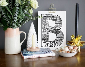 May include: Black and white illustration of the city of Bristol, England, with the city name written in large letters below the illustration. The illustration is framed and hanging on a gray wall. There are decorative items on a wooden table in front of the illustration, including a vase with flowers, a candle, a bowl of ornaments, and a star.