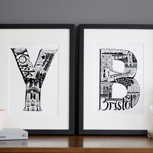 May include: Two black framed prints of the letters Y and B, each letter is made up of a black and white illustration of a city skyline. The Y print has the word "York" at the bottom and the B print has the word "Bristol" at the bottom.