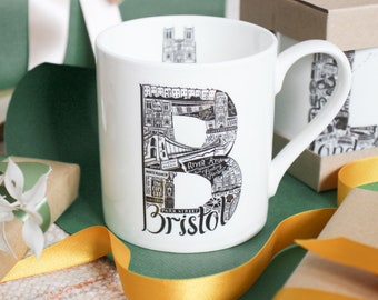 May include: White ceramic mug featuring a black and white illustration of Bristol landmarks within a large letter "B". The mug includes the word "Bristol" at the bottom. The mug is surrounded by gift boxes and ribbons.