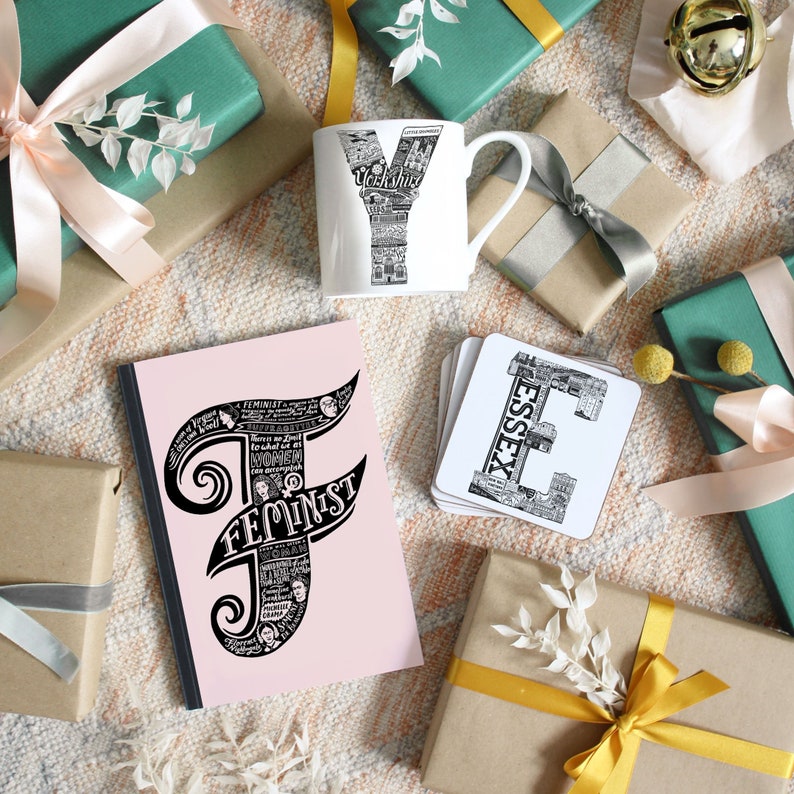 May include: A collection of gifts including a white mug with a black and white Yorkshire design, a pink notebook with the word "FEMINIST" in black, and coasters with an "ESSEX" design. The gifts are surrounded by wrapped presents.
