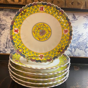 May include: A stack of six round porcelain plates with scalloped edges. Each plate features a bright yellow border decorated with intricate red, blue, and green patterns, including four red Chinese characters and a central floral medallion design.