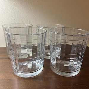 May include: A set of four clear glass tumblers, each featuring a textured grid pattern of raised squares on the exterior surface. The glasses have a cylindrical shape with a smooth rim and a thick, solid base, providing a uniform appearance.