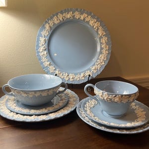 May include: A light blue ceramic dinnerware set featuring white embossed grape vine relief patterns. The collection includes a large dinner plate, a two-handled soup bowl with a matching saucer, and a tea cup with a matching saucer.