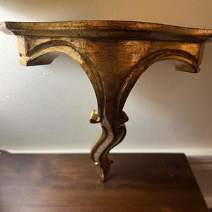 May include: A wall-mounted shelf featuring a gold-toned finish with a weathered, antique appearance. The shelf has a flat top surface and a decorative, curved support bracket that extends downward, showcasing intricate carved details.