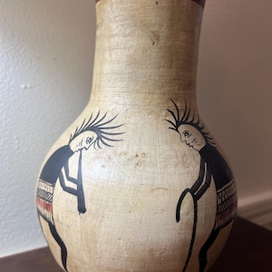 May include: A tan ceramic vase with a narrow neck and a rounded body. The vase features a dark red band around the rim and black painted figures of two individuals with spiky hair. One figure plays a flute while the other stands nearby.