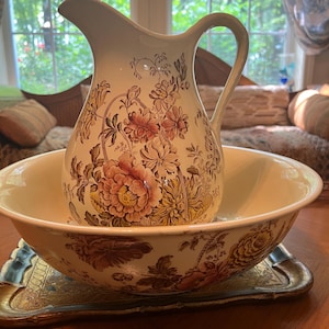 May include: Cream-colored pitcher and basin set with a floral pattern in shades of brown, pink, and yellow. The pitcher has a curved handle and spout. The basin sits on a decorative tray, creating a vintage aesthetic.