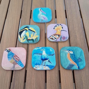Puede incluir: Una colección de seis posavasos cuadrados, cada uno pintado a mano con un diseño de ave diferente. Las aves incluyen un pelícano, tucanes, una abubilla, una codorniz, una gaviota y un martín pescador. Los posavasos están colocados sobre una superficie de madera.