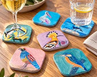 Può includere: Un set di sei sottobicchieri quadrati con illustrazioni di uccelli dipinte a mano, tra cui un tucano, un pellicano, un'upupa, una quaglia e un martin pescatore, disposti su una superficie in legno con un calice e un bicchiere d'acqua.