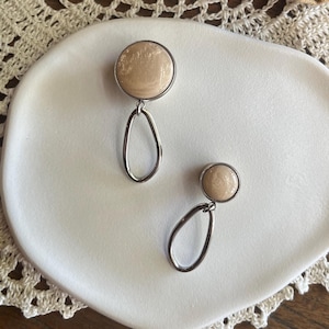 Op de afbeelding: Een paar oorbellen met een licht perzik kleurige cirkelvormige stenen top en een zilveren druppelvormige hanger. De oorbellen worden gepresenteerd op een witte, organisch gevormde plaat, tegen een achtergrond van een kanten kleedje en een houten oppervlak.