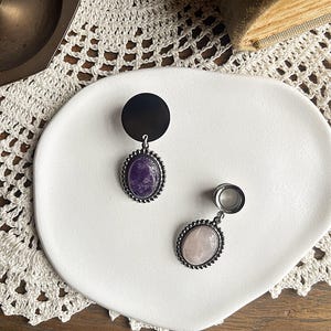 Op de afbeelding: Een paar zilverkleurige oortunnels met ovale edelsteenhangers. Eén heeft een paarse steen, de andere een roze steen. De tunnels hebben een kralenrand en een zwarte schijf. De sieraden worden gepresenteerd op een wit bord.