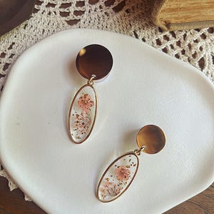 May include: A pair of gold-toned earrings. Each earring features a circular top and an oval pendant. The pendants are clear, showcasing dried pink flowers and gold flecks. The earrings rest on a white plate.
