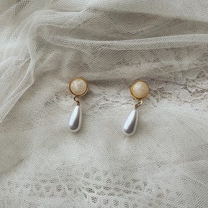 Op de afbeelding: Een paar goudkleurige oorknopjes met een ronde, getextureerde crèmekleurige cabochon aan de bovenkant en een bungelende, gladde witte druppelvormige kunstparel hanger bevestigd aan een kleine goudkleurige metalen ring.