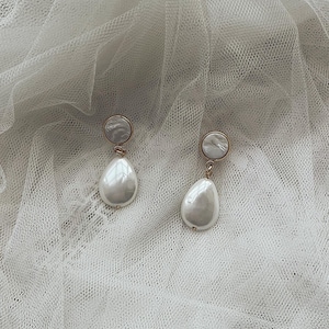 May include: A pair of drop earrings featuring a circular stud top with a pearlescent finish and a dangling, smooth, teardrop-shaped white pearl-like pendant. The components are connected by small gold-toned metal rings, creating a classic design.