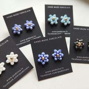 May include: Five pairs of flower-shaped porcelain stud earrings displayed on black cards labeled HAND MADE PORCELAIN. The earrings come in white, light blue, periwinkle, dark blue, and dark gray, each featuring a small gold-colored center bead.