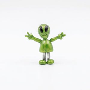 May include: A green alien figurine with a smiling face and outstretched arms. The alien has large black eyes and is mounted on a silver spring base. The figurine's feet are also green.