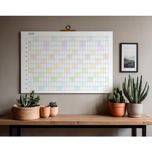 May include: A large white 2026 year planner with colourful blocks hangs on a neutral wall. Below, a wooden desk displays potted plants, including succulents and cacti. A framed black and white landscape photo is also on the wall.