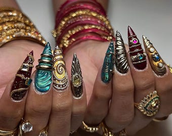 May include: A close-up of elaborately decorated long nails. The nails feature a variety of designs, including gold, teal, and burgundy accents. The nails are adorned with jewels and intricate patterns. Gold rings and bracelets are also visible.
